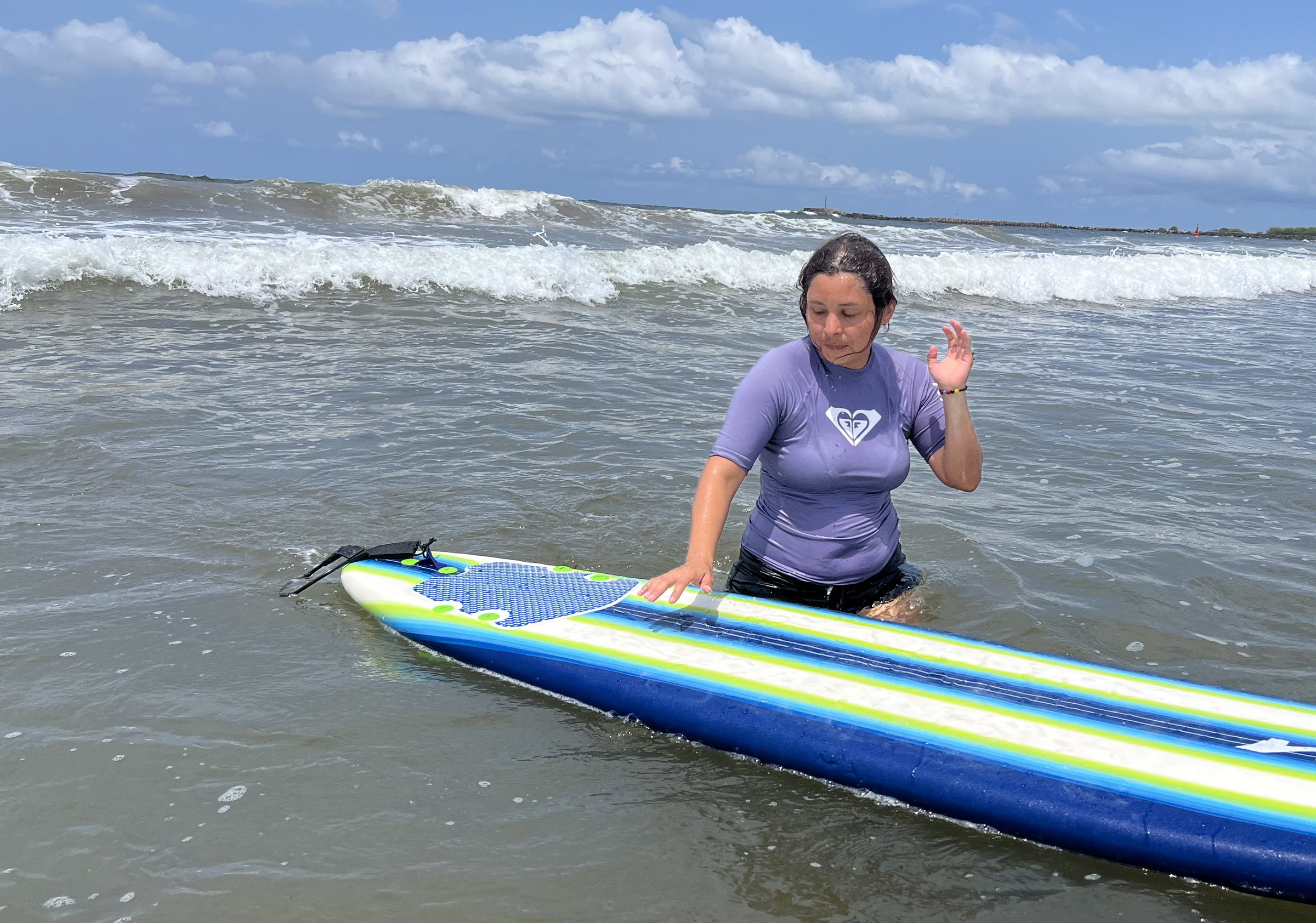 Alumna de pie en el agua junto a su tabla, saludando a la cámara