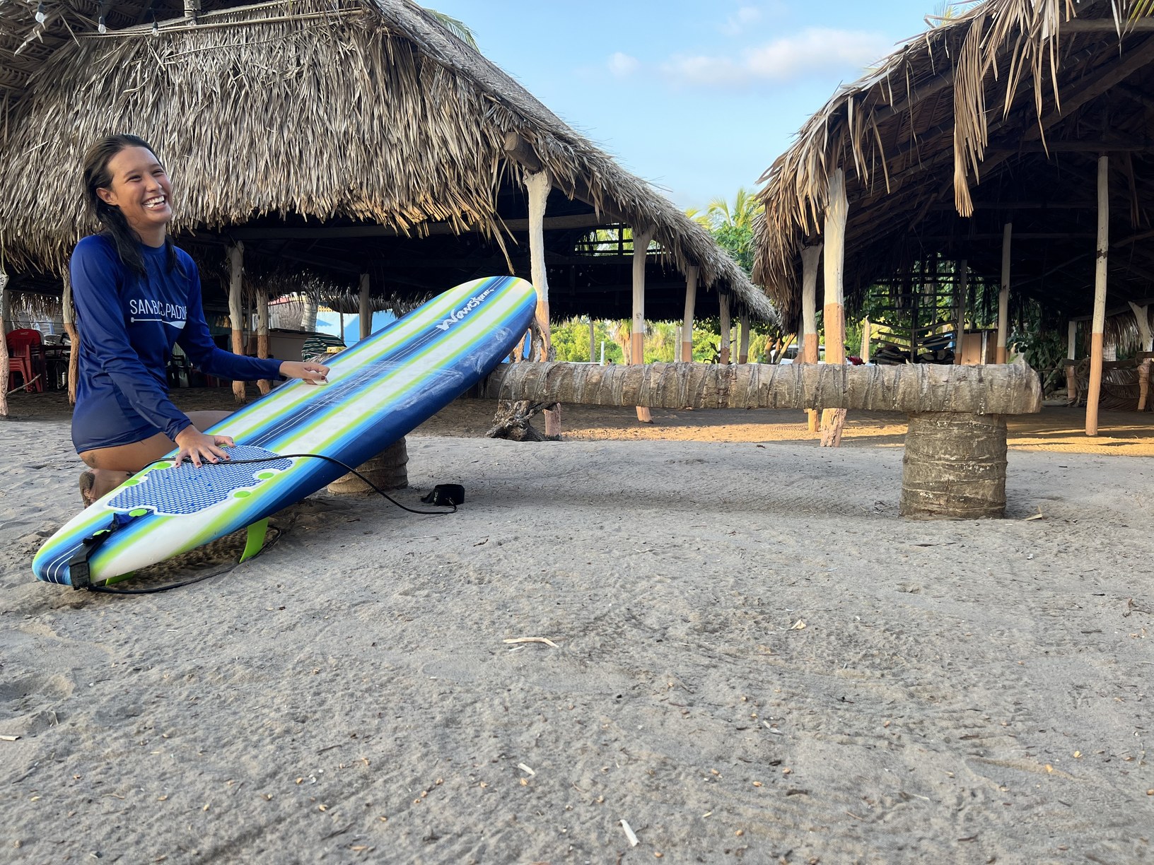 Alumna sonriendo junto a su tabla bajo las palapas, antes de entrar al agua