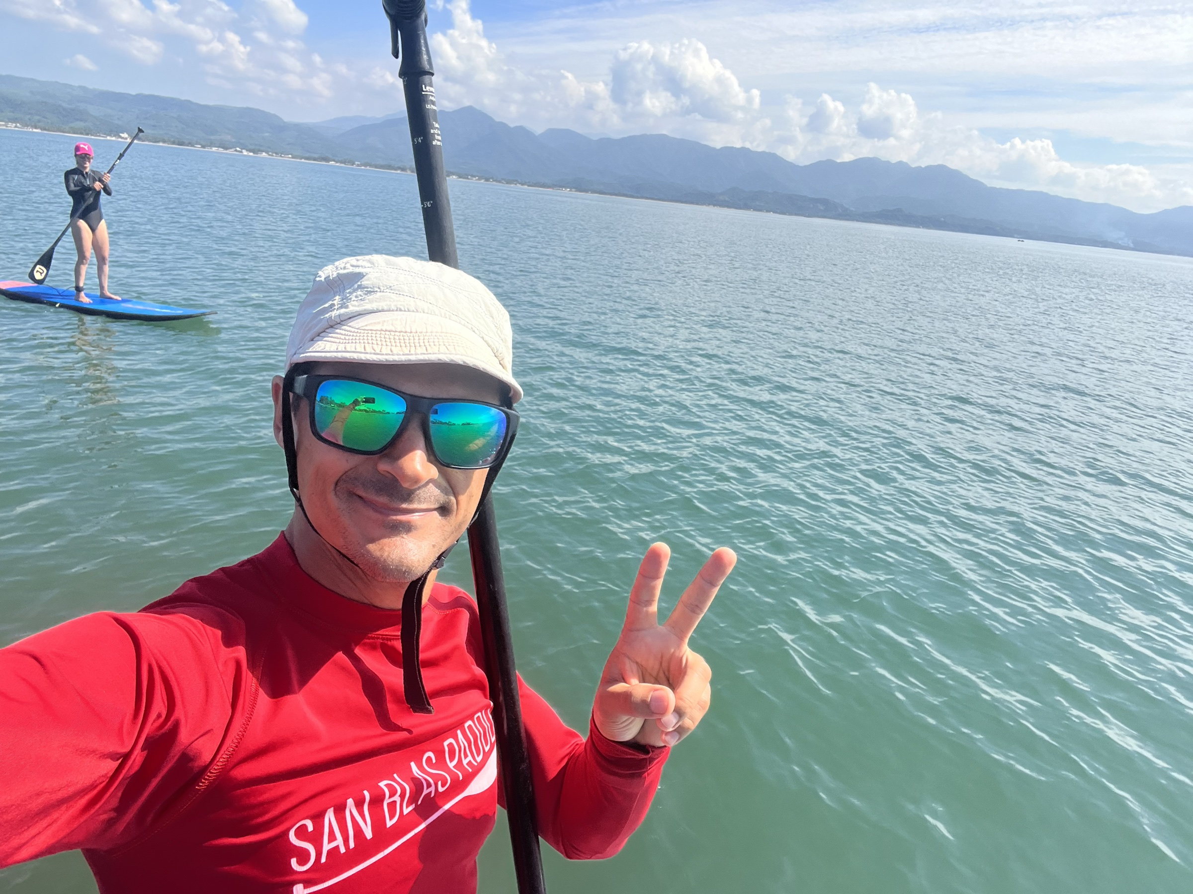 Instructor de paddleboard en el agua junto a una alumna que rema de pie sobre su tabla, con la sierra al fondo