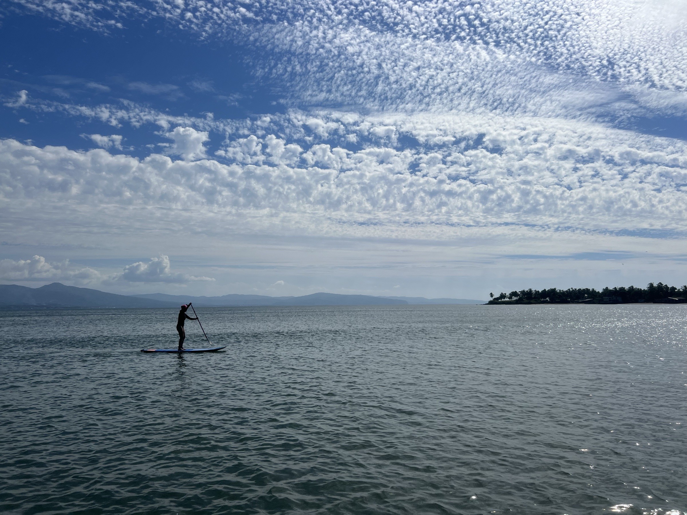 Persona remando de pie sobre un paddleboard en el agua plana de Las Islitas, con la sierra al fondo