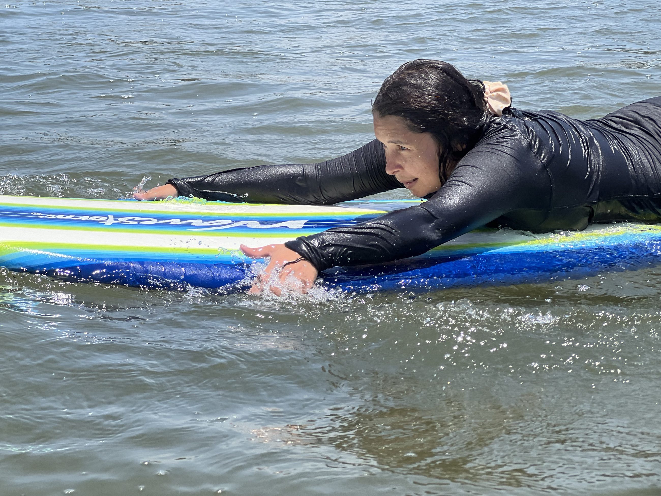 Alumna con traje de neopreno negro remando acostada sobre una tabla grande de principiante, en agua tranquila