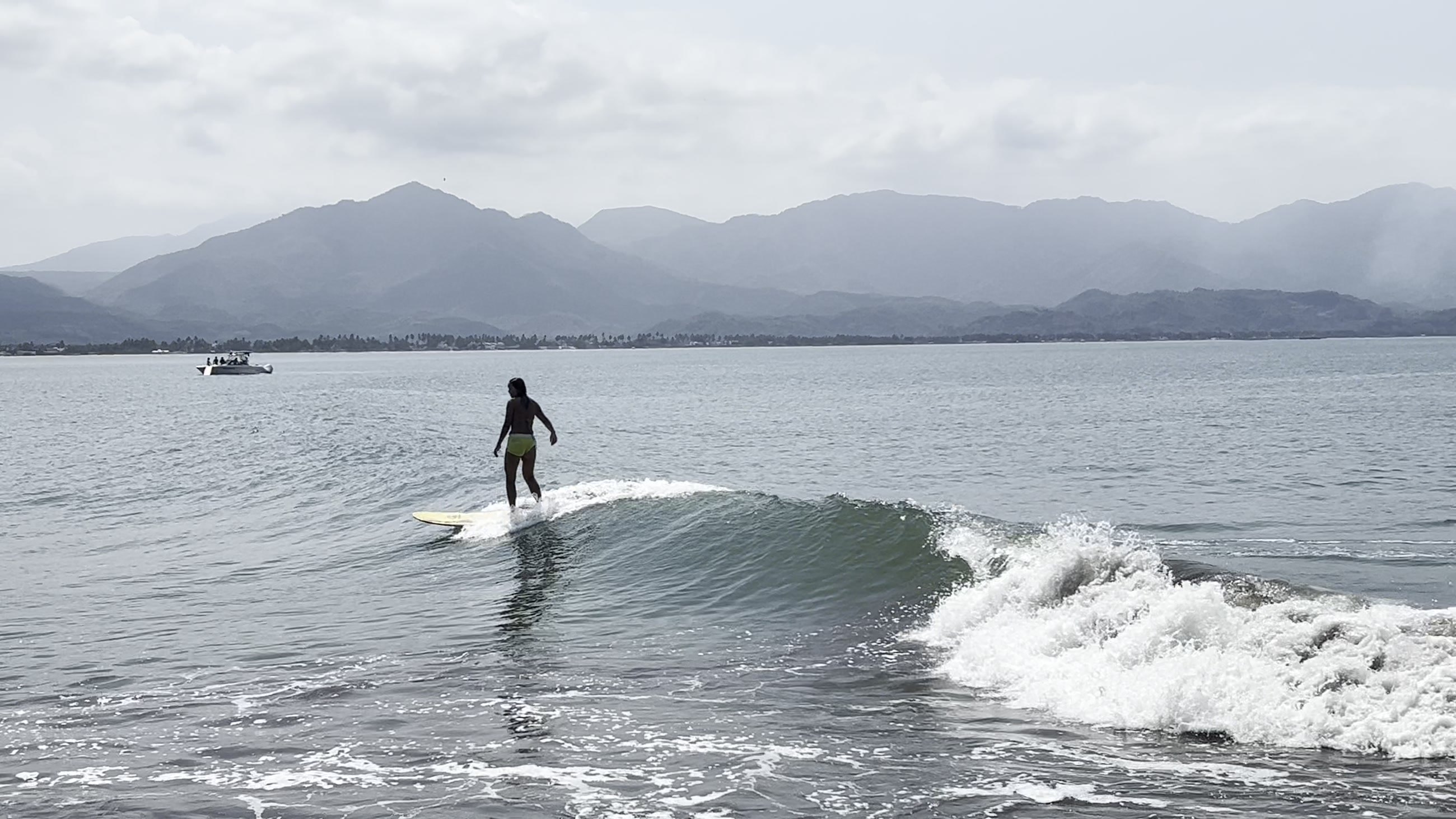 Surfista recorriendo de pie la pared verde de una ola bien formada, con la sierra y una panga al fondo