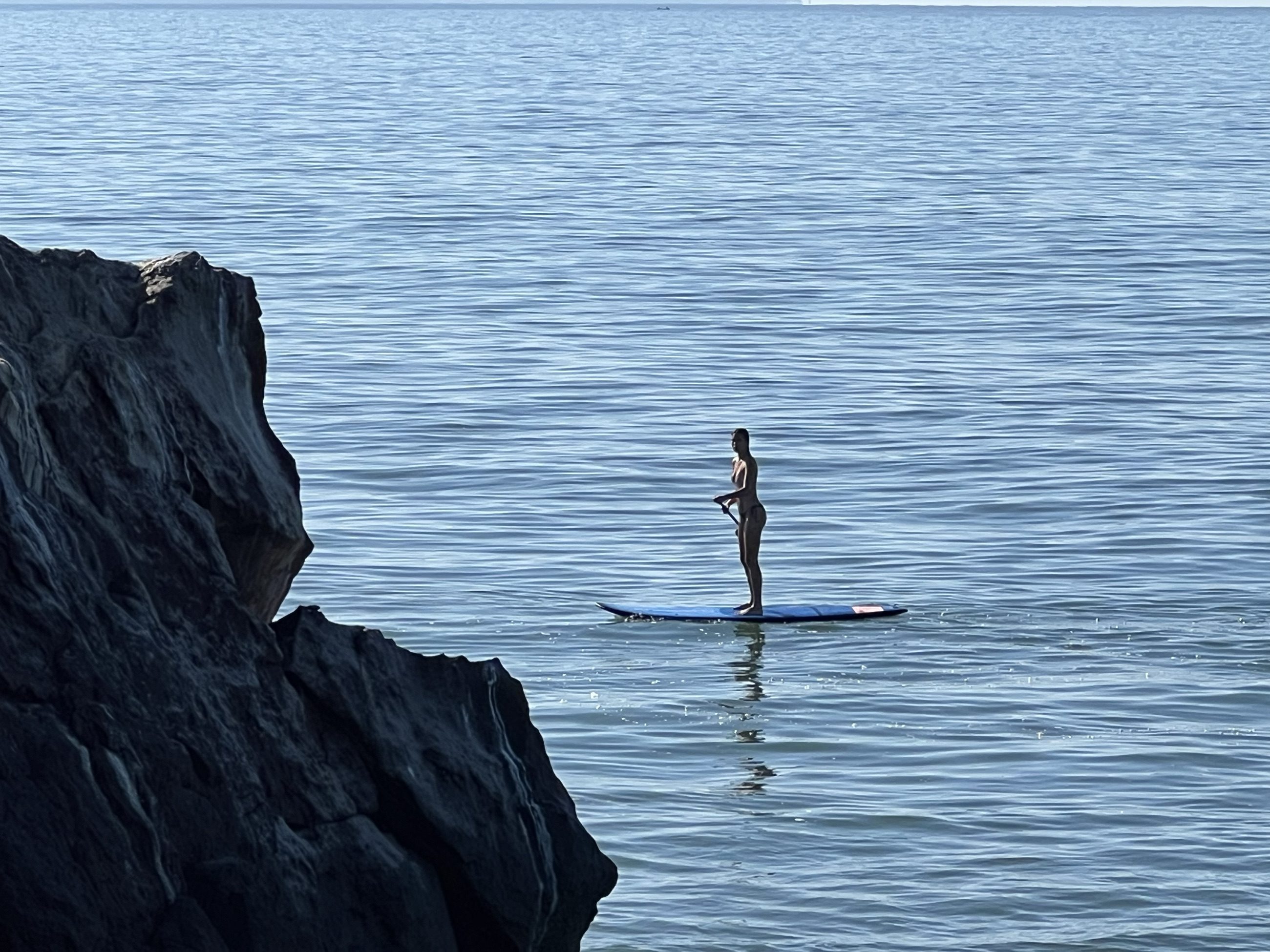 Persona remando de pie sobre un paddleboard en aguas tranquilas, enmarcada por una roca oscura en primer plano